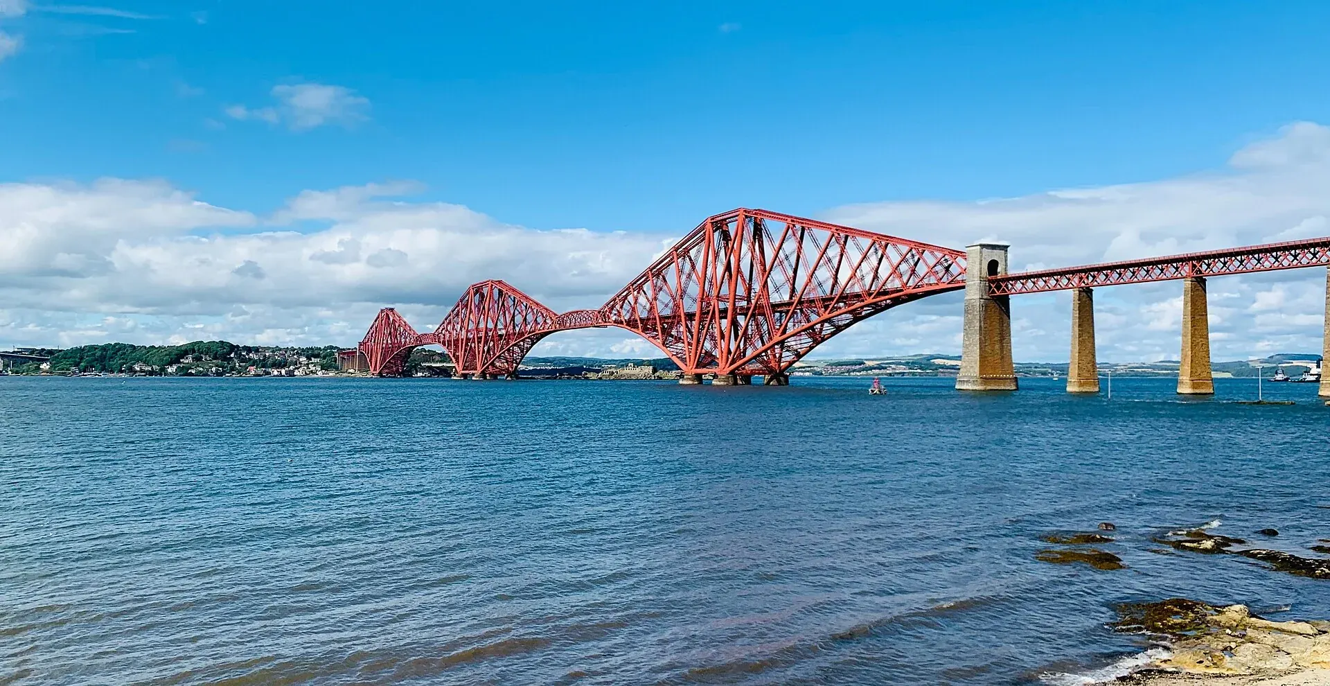 The red steel Forth Bridge spanning blue water beneath a bright sky, viewed from the south shore