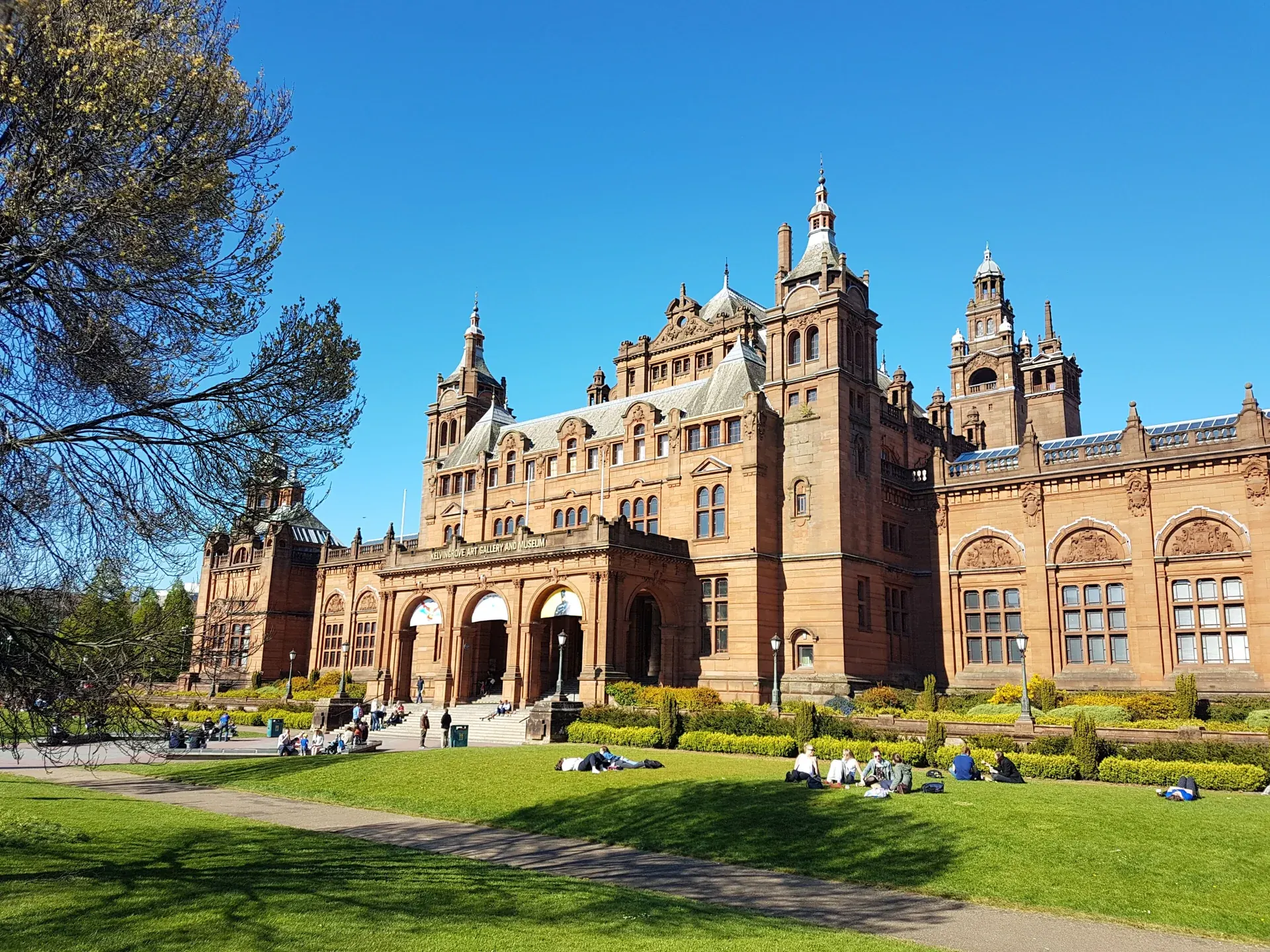 Red sandstone Kelvingrove Art Gallery and Museum beside green lawns beneath a blue sky in Glasgow
